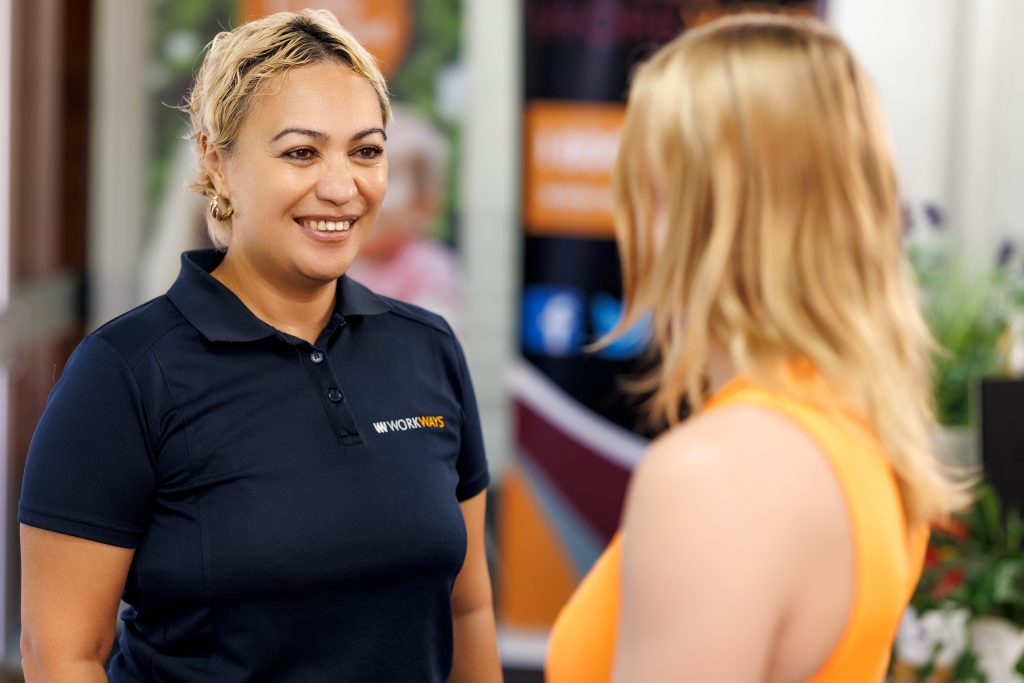 Woman smiling wearing navy blue uniform t-shirt with Workways logo in white, talking to female customer in orange singlet